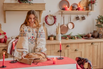 A woman wearing a traditional embroidered dress is placing a freshly baked loaf of bread on a table covered with a red cloth. The room is decorated for the holiday season with evergreen arrangements and red candles. Copper pans and other utensils hang on the wall, adding a rustic charm.