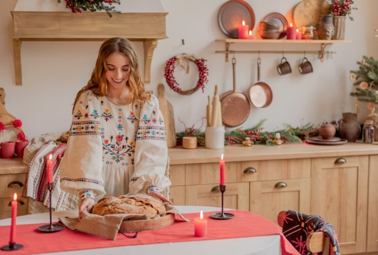 A woman wearing a traditional embroidered dress is placing a freshly baked loaf of bread on a table covered with a red cloth. The room is decorated for the holiday season with evergreen arrangements and red candles. Copper pans and other utensils hang on the wall, adding a rustic charm.