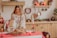 A woman wearing a traditional embroidered dress is placing a freshly baked loaf of bread on a table covered with a red cloth. The room is decorated for the holiday season with evergreen arrangements and red candles. Copper pans and other utensils hang on the wall, adding a rustic charm.