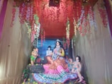 A colorful and ornate scene with three statues inside a decorated enclosure. The central figure is a female deity in vibrant attire, seated with flowing garments, accompanied by two smaller statues of children or deities. Flower garlands in red, pink, and white hang from above, adding a vibrant touch.