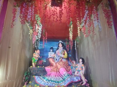 A colorful and ornate scene with three statues inside a decorated enclosure. The central figure is a female deity in vibrant attire, seated with flowing garments, accompanied by two smaller statues of children or deities. Flower garlands in red, pink, and white hang from above, adding a vibrant touch.