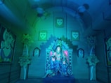 A detailed and colorful interior with a statue of a deity seated on a throne in the center, surrounded by decorative elements like flowers and paintings. The walls and ceiling are adorned with faces and abstract designs, creating a mystical, temple-like atmosphere.