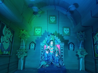 A detailed and colorful interior with a statue of a deity seated on a throne in the center, surrounded by decorative elements like flowers and paintings. The walls and ceiling are adorned with faces and abstract designs, creating a mystical, temple-like atmosphere.