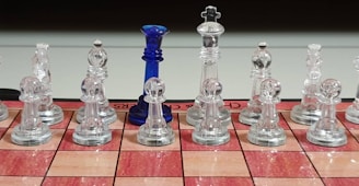 A chessboard set with transparent glass pieces is arranged for a game. Among the clear chess pieces, one distinct blue piece stands out. The chessboard has a red and tan checkered pattern, and the pieces are neatly aligned in two rows.