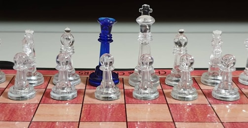 A chessboard set with transparent glass pieces is arranged for a game. Among the clear chess pieces, one distinct blue piece stands out. The chessboard has a red and tan checkered pattern, and the pieces are neatly aligned in two rows.