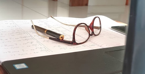A pair of eyeglasses with brown frames rests on top of an open notebook filled with handwritten notes. A pen with a gold and black design lies near the glasses. Part of a laptop and a mobile phone are partially visible in the foreground and background, respectively.