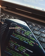 A close-up view of a laptop keyboard with a smartphone resting on top. The smartphone screen displays colorful lines of programming code, reflected prominently against the keyboard. The laptop screen also shows some text or code in the background.
