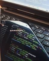 A close-up view of a laptop keyboard with a smartphone resting on top. The smartphone screen displays colorful lines of programming code, reflected prominently against the keyboard. The laptop screen also shows some text or code in the background.