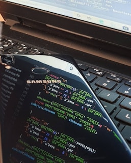 A close-up view of a laptop keyboard with a smartphone resting on top. The smartphone screen displays colorful lines of programming code, reflected prominently against the keyboard. The laptop screen also shows some text or code in the background.