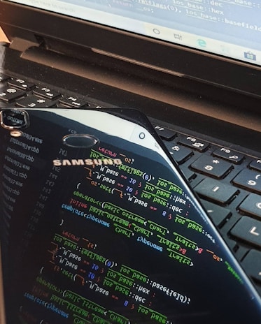 A close-up view of a laptop keyboard with a smartphone resting on top. The smartphone screen displays colorful lines of programming code, reflected prominently against the keyboard. The laptop screen also shows some text or code in the background.