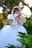 A young girl with blonde hair is wearing a white dress with gold embroidery, standing amidst lush green foliage and holding white calla lilies. The background is softly blurred with sunlight filtering through trees, creating a serene and dreamy atmosphere.