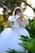 A young girl with blonde hair is wearing a white dress with gold embroidery, standing amidst lush green foliage and holding white calla lilies. The background is softly blurred with sunlight filtering through trees, creating a serene and dreamy atmosphere.