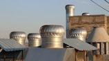 Several metallic ventilation turbines are installed on the rooftop of a building, surrounded by other metal structures. The roof has a mix of textures with corrugated and plain surfaces. The turbines have a shiny, silver appearance and are catching the light of the setting sun. The sky in the background is clear with a soft, warm light.