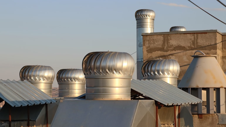 Several metallic ventilation turbines are installed on the rooftop of a building, surrounded by other metal structures. The roof has a mix of textures with corrugated and plain surfaces. The turbines have a shiny, silver appearance and are catching the light of the setting sun. The sky in the background is clear with a soft, warm light.