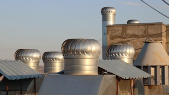 Several metallic ventilation turbines are installed on the rooftop of a building, surrounded by other metal structures. The roof has a mix of textures with corrugated and plain surfaces. The turbines have a shiny, silver appearance and are catching the light of the setting sun. The sky in the background is clear with a soft, warm light.
