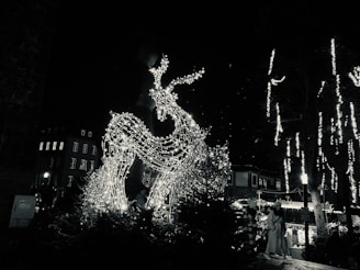 A large, illuminated reindeer sculpture is adorned with white string lights, creating a festive display. The scene is set in a dimly lit outdoor area with other illuminated decorations and a backdrop of buildings. People are visible nearby, possibly enjoying a holiday market.