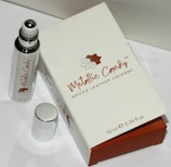 A small bottle of cologne labeled 'Metallic Candy' with a spiced leather scent is positioned next to its packaging box. The bottle has a metallic rollerball applicator and is placed alongside a shiny silver cap. The box is predominantly white with brown accents and displays the brand name and perfume details.