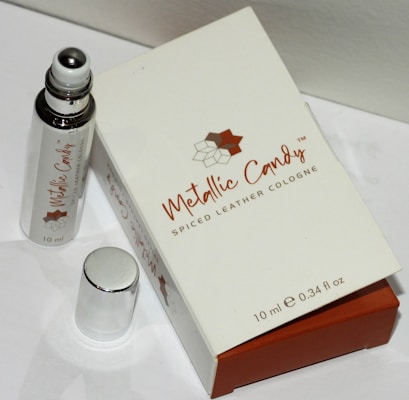 A small bottle of cologne labeled 'Metallic Candy' with a spiced leather scent is positioned next to its packaging box. The bottle has a metallic rollerball applicator and is placed alongside a shiny silver cap. The box is predominantly white with brown accents and displays the brand name and perfume details.
