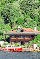 A large, modern house with a wooden facade is situated by the water's edge, surrounded by lush, green forested hills. In front of the house, a sleek red and white boat is docked at a small pier. The scene is bright and sunny, suggesting a peaceful, leisurely environment.