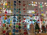 A colorful display of various hanging paper decorations, including hearts, circles, butterflies, stars, and owls. The decorations are suspended from the ceiling in multiple rows and layers, creating a vibrant and festive atmosphere. The background features a store setting with windows and shelves filled with various items.