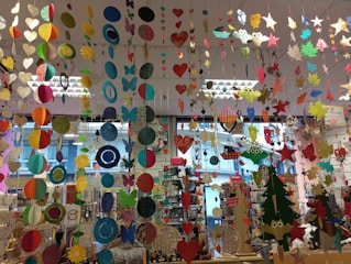 A colorful display of various hanging paper decorations, including hearts, circles, butterflies, stars, and owls. The decorations are suspended from the ceiling in multiple rows and layers, creating a vibrant and festive atmosphere. The background features a store setting with windows and shelves filled with various items.