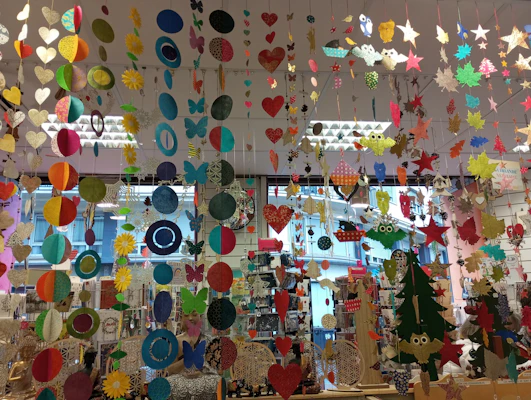 A colorful display of various hanging paper decorations, including hearts, circles, butterflies, stars, and owls. The decorations are suspended from the ceiling in multiple rows and layers, creating a vibrant and festive atmosphere. The background features a store setting with windows and shelves filled with various items.