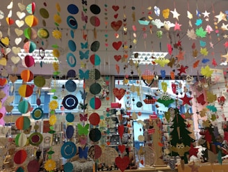 A colorful display of various hanging paper decorations, including hearts, circles, butterflies, stars, and owls. The decorations are suspended from the ceiling in multiple rows and layers, creating a vibrant and festive atmosphere. The background features a store setting with windows and shelves filled with various items.