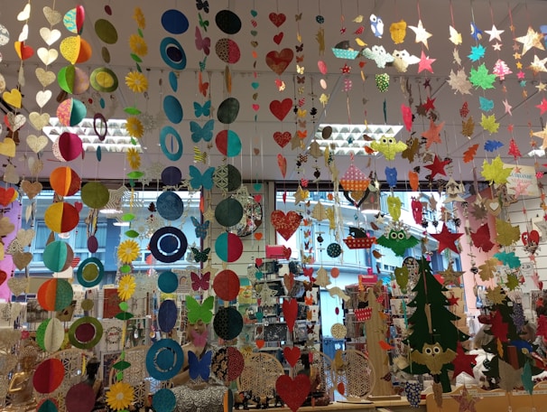 A colorful display of various hanging paper decorations, including hearts, circles, butterflies, stars, and owls. The decorations are suspended from the ceiling in multiple rows and layers, creating a vibrant and festive atmosphere. The background features a store setting with windows and shelves filled with various items.