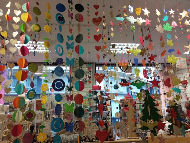 A colorful display of various hanging paper decorations, including hearts, circles, butterflies, stars, and owls. The decorations are suspended from the ceiling in multiple rows and layers, creating a vibrant and festive atmosphere. The background features a store setting with windows and shelves filled with various items.