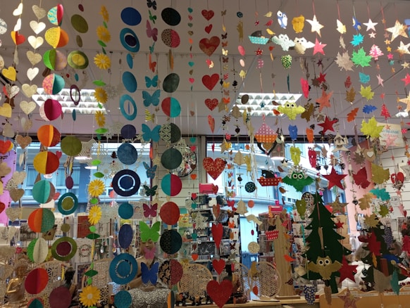 A colorful display of various hanging paper decorations, including hearts, circles, butterflies, stars, and owls. The decorations are suspended from the ceiling in multiple rows and layers, creating a vibrant and festive atmosphere. The background features a store setting with windows and shelves filled with various items.