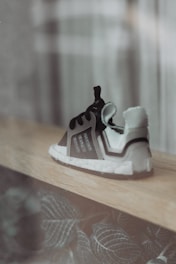 A single sneaker is placed on a wooden surface, featuring a modern design with a combination of black and white colors. The background includes soft-focus patterns and leaves with a muted green hue.