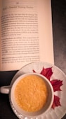 A book open to a page titled 'Build a Powerful Morning Routine' is situated on a table beside a white cup filled with a frothy beverage, likely coffee or tea, on a saucer. Beside the cup, there are two autumn leaves, providing a seasonal touch to the scene.