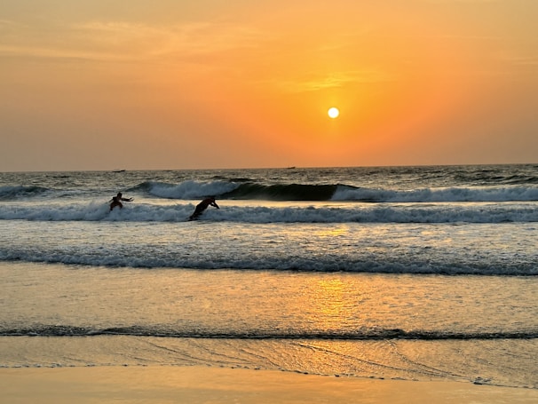 Surfers catching gentle waves under a golden sunset on a Moroccan beach.