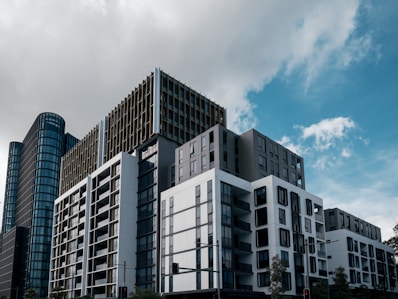 A modern urban landscape featuring several high-rise buildings with contemporary architecture under a partly cloudy sky. The structures are composed of glass, concrete, and metal elements, and exhibit a sleek, geometric design.