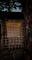 A yellow sign with red lettering lists food items such as nasi goreng, nasi mawut, mie goreng, and mie kuah along with their price. The sign is attached to a wooden frame structure and there is a large tree on the right side.