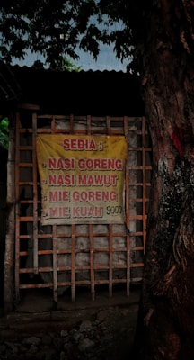 A yellow sign with red lettering lists food items such as nasi goreng, nasi mawut, mie goreng, and mie kuah along with their price. The sign is attached to a wooden frame structure and there is a large tree on the right side.