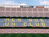 A football stadium with empty seats forming the phrase 'MES QUE UN CLUB' in large yellow letters on a blue background. The stands are predominantly empty with alternating rows of red and blue seating. Advertising banners are visible along the top edges of the upper tier.