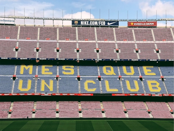 A football stadium with empty seats forming the phrase 'MES QUE UN CLUB' in large yellow letters on a blue background. The stands are predominantly empty with alternating rows of red and blue seating. Advertising banners are visible along the top edges of the upper tier.