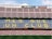 A football stadium with empty seats forming the phrase 'MES QUE UN CLUB' in large yellow letters on a blue background. The stands are predominantly empty with alternating rows of red and blue seating. Advertising banners are visible along the top edges of the upper tier.