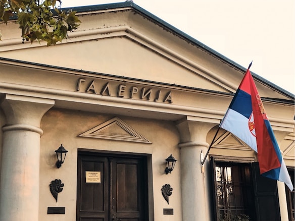 The image features the entrance of a neoclassical building with a triangular pediment and two columns flanking a wooden double door. Above the entrance, there is an inscription in Cyrillic script. A Serbian flag is prominently displayed, attached to the right side of the building. Two wall-mounted lanterns and decorative carvings are also visible.