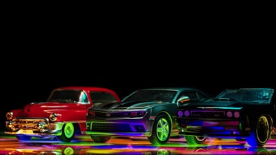 A row of three toy cars is placed in a line against a black background. The cars include a red classic model on the left, a green-highlighted sports car in the center, and a dark, sleek car with colorful reflections on the right. The ground reflects vibrant, colorful lights which add a dynamic and lively feel to the scene.