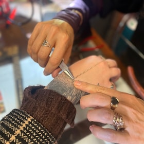 Two hands are working with tools on a chain held over a tattooed wrist. The hands have rings and are partially covered by warm clothing. The setting appears to be a workshop with a focus on jewelry or accessories.