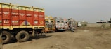 A row of vibrantly decorated trucks is parked on a dirt lot. The trucks feature intricate patterns and bright colors, typical of traditional artistic styles, including motifs like flowers and abstract designs. A motorbike is parked among the trucks, and the background shows a few buildings and scattered foliage.