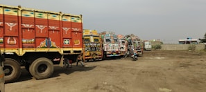 A row of vibrantly decorated trucks is parked on a dirt lot. The trucks feature intricate patterns and bright colors, typical of traditional artistic styles, including motifs like flowers and abstract designs. A motorbike is parked among the trucks, and the background shows a few buildings and scattered foliage.