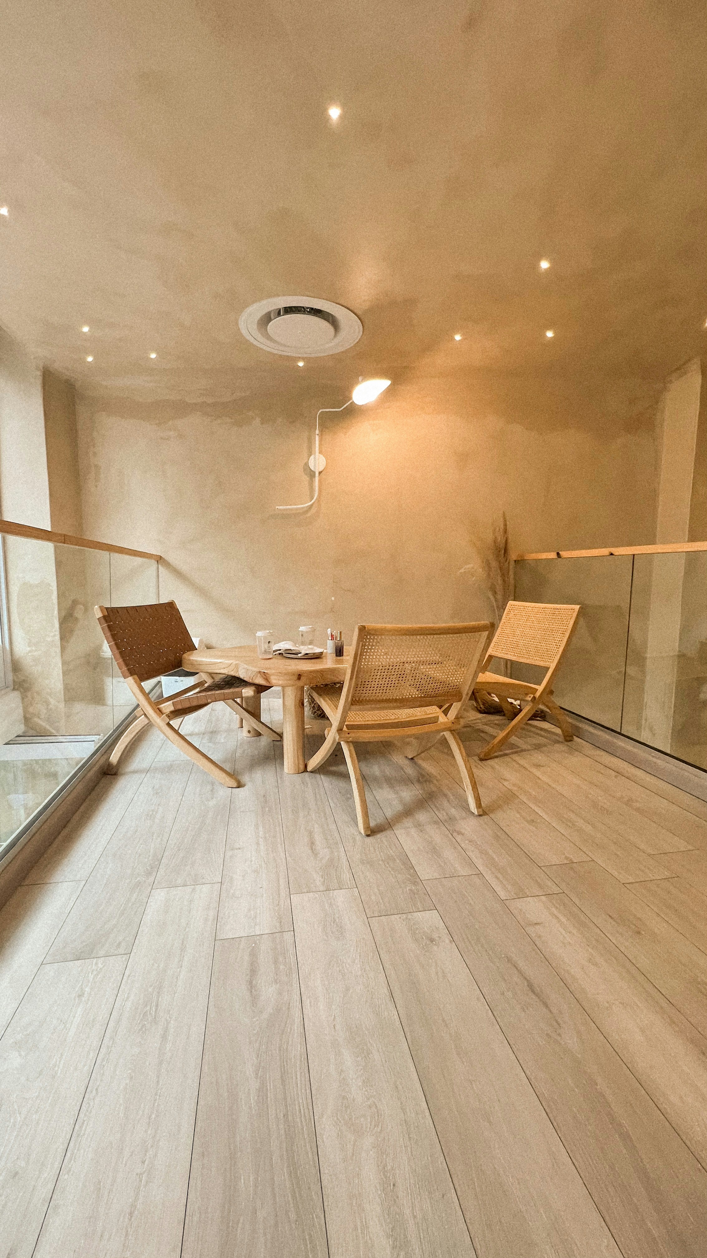 A minimalist and cozy room features wooden flooring, a small round wooden table surrounded by three woven chairs. The walls and ceiling are painted in a warm beige color, with recessed lighting that adds a soft glow. There is a modern ventilation unit on the ceiling, and the space is bordered by glass panels.