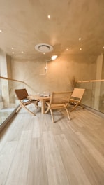A minimalist and cozy room features wooden flooring, a small round wooden table surrounded by three woven chairs. The walls and ceiling are painted in a warm beige color, with recessed lighting that adds a soft glow. There is a modern ventilation unit on the ceiling, and the space is bordered by glass panels.