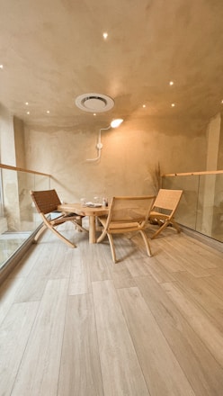 A minimalist and cozy room features wooden flooring, a small round wooden table surrounded by three woven chairs. The walls and ceiling are painted in a warm beige color, with recessed lighting that adds a soft glow. There is a modern ventilation unit on the ceiling, and the space is bordered by glass panels.