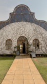 A rustic building with a curved roof partially covered in thin vines. Two symmetrical windows and a wooden door are centrally placed on the white facade. A stone pathway leads to the entrance, flanked by grass and small hedges. Chalkboards with handwritten text are positioned on either side of the doorway.