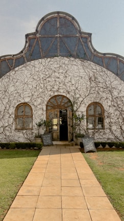 A rustic building with a curved roof partially covered in thin vines. Two symmetrical windows and a wooden door are centrally placed on the white facade. A stone pathway leads to the entrance, flanked by grass and small hedges. Chalkboards with handwritten text are positioned on either side of the doorway.