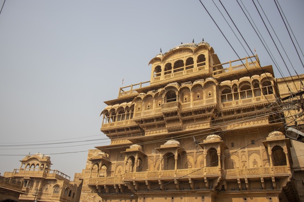 An intricately designed sandstone building with multiple floors and arched windows, featuring detailed carvings and traditional Indian architectural elements. Overhead electrical wires are visible, crisscrossing in front of the structure, and the sky is clear and light blue.